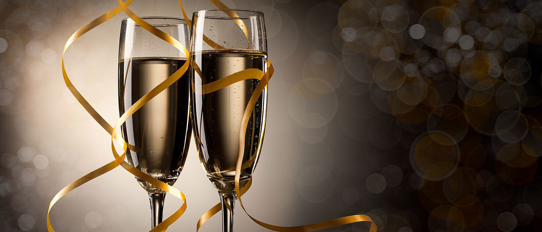 Zwei gefüllte Sektgläser stehen nebeneinander vor einem dunklen, festlichen Hintergrund. Goldene Luftschlangen winden sich elegant um die Gläser. Die stimmungsvolle Beleuchtung erzeugt eine edle, feierliche Atmosphäre – passend zu einer Silvesterfeier und weiteren Feieranlässen.