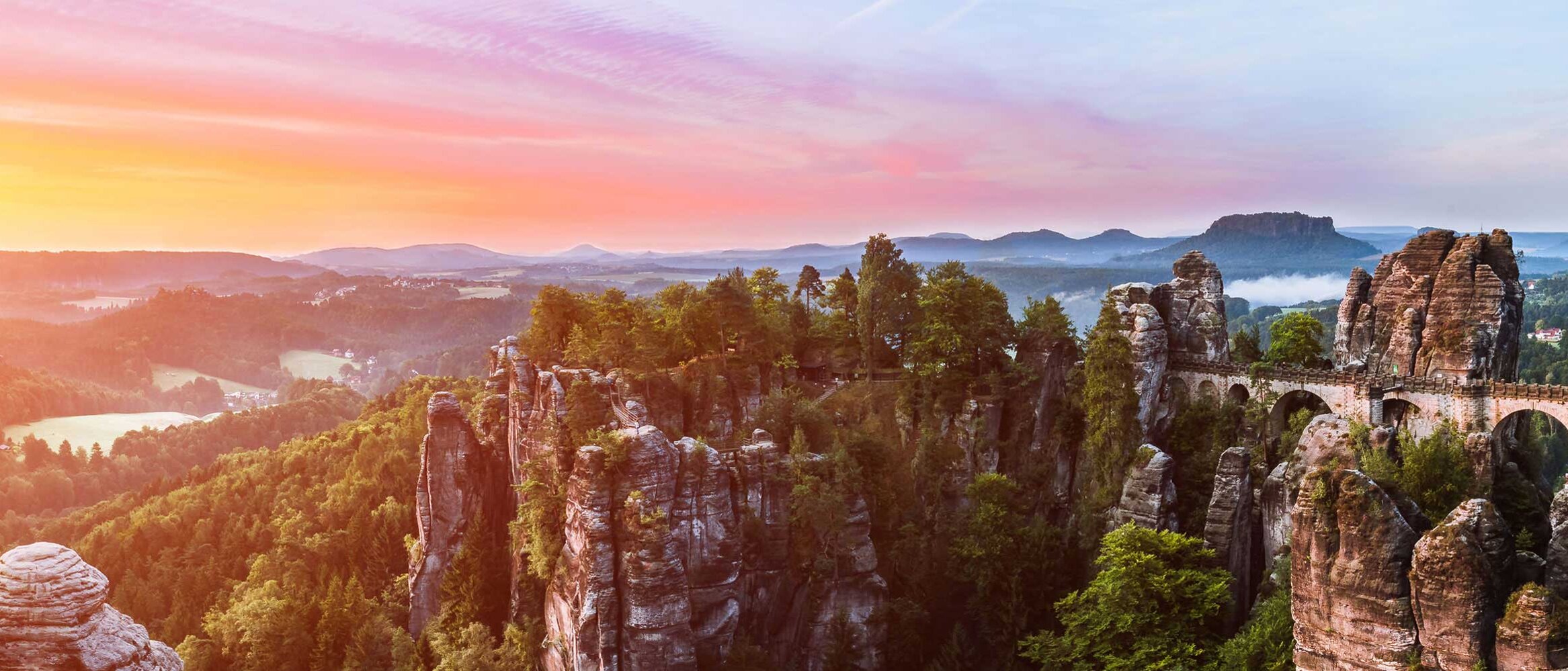 Panoramaaufnahme der markanten Sandsteinfelsen und bewaldeten Hügel des Elbsandsteingebirges bei Sonnenaufgang, mit Blick auf die Umgebung von Pirna – Naturlandschaft am Aktiv Sporthotel.