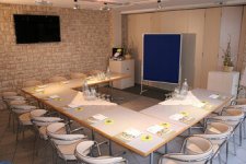 Moderner Seminarraum im Material-Mix: Vor einer markanten Natursteinwand mit großem Präsentations-Bildschirm ist eine U-förmige Tafel aufgebaut. Die hellen Polsterstühle mit Chromgestell wirken leicht und modern. Arbeitsunterlagen, Getränke und eine blaue Pinnwand stehen für das nächste Meeting bereit.