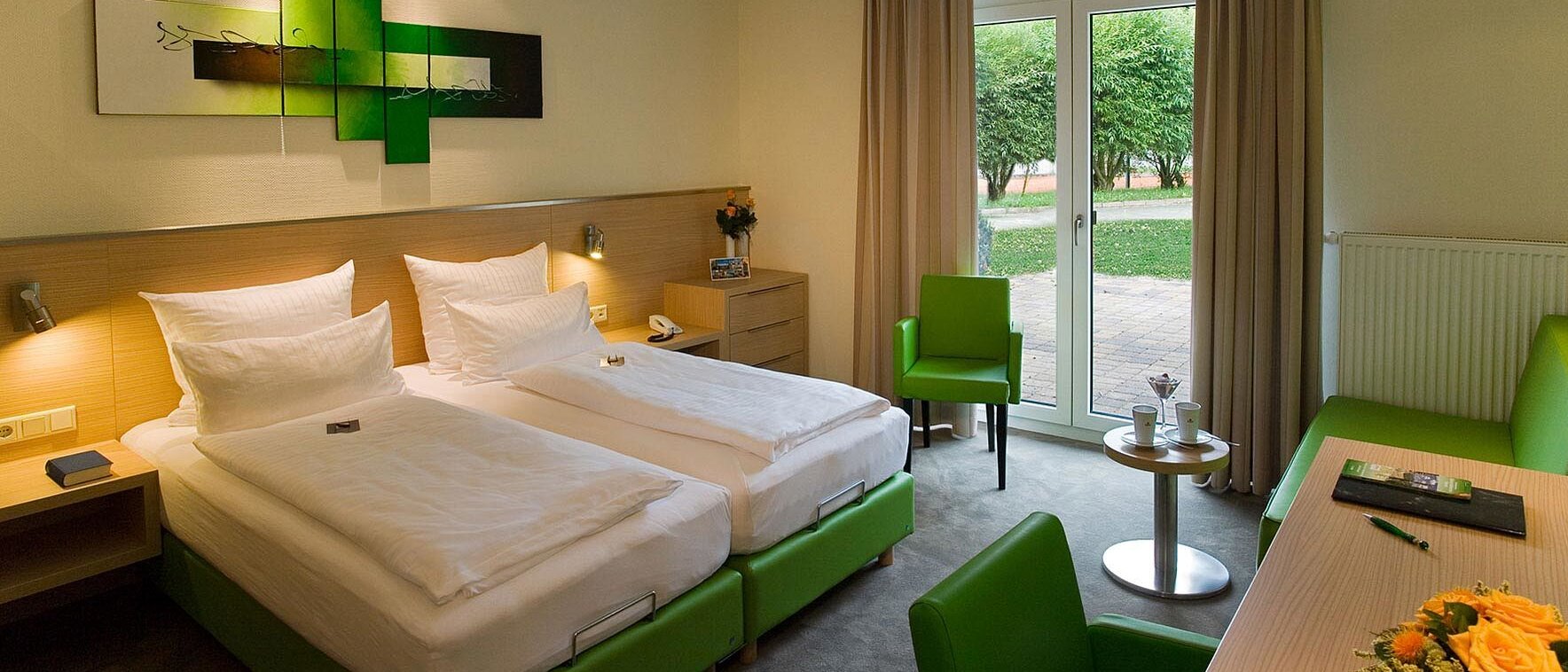 Das Bild zeigt ein modernes, freundlich eingerichtetes Hotelzimmer im aktiv Sporthotel Pirna in der Sächsischen Schweiz. Der Raum wirkt hell und einladend, da viel Tageslicht durch eine große Terrassentür einfällt, die sich ebenerdig öffnen lässt und einen direkten Zugang ins Freie bietet – ein Pluspunkt für Barrierefreiheit.  Im Zentrum des Zimmers stehen zwei niedrige, gut zugängliche Einzelbetten, die nebeneinander angeordnet sind. Sie haben weiße Bettwäsche und breite, gepolsterte Kopfteile. Über den Betten hängt ein modernes, abstraktes Wandbild in Grün- und Schwarztönen.  Rechts im Bild befindet sich ein Schreibtisch mit einem Stuhl, auf dem eine Tischlampe und Informationsmaterial liegen. Alle Möbel sind so platziert, dass sich breite Wege ergeben, was eine gute Beweglichkeit im Raum ermöglicht.  Links und rechts im Zimmer stehen zusätzliche Stühle mit Armlehnen, die für Menschen mit eingeschränkter Mobilität hilfreich sein können. Die Farbgestaltung ist ruhig und natürlich: helle Wände, grüne Akzente an Stühlen, Betten und Dekoration.  Das Zimmer wirkt insgesamt barrierearm, gut beleuchtet und strukturiert, sodass es sich für Gäste mit unterschiedlichen Bedürfnissen eignet.
