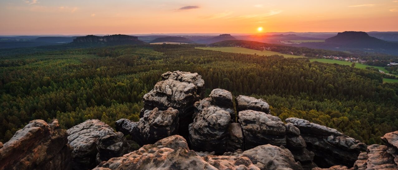 Blick von einem Felsplateau über die weitläufige Landschaft des Elbsandsteingebirges. Im Vordergrund liegen helle, kantige Felsen, dahinter erstrecken sich Hügel, Wälder und Tafelberge in warmen Herbsttönen. Die Sonne steht tief am Horizont und taucht die Szenerie in goldenes Licht – ein stimmungsvolles Motiv für ein Herbst-Special.