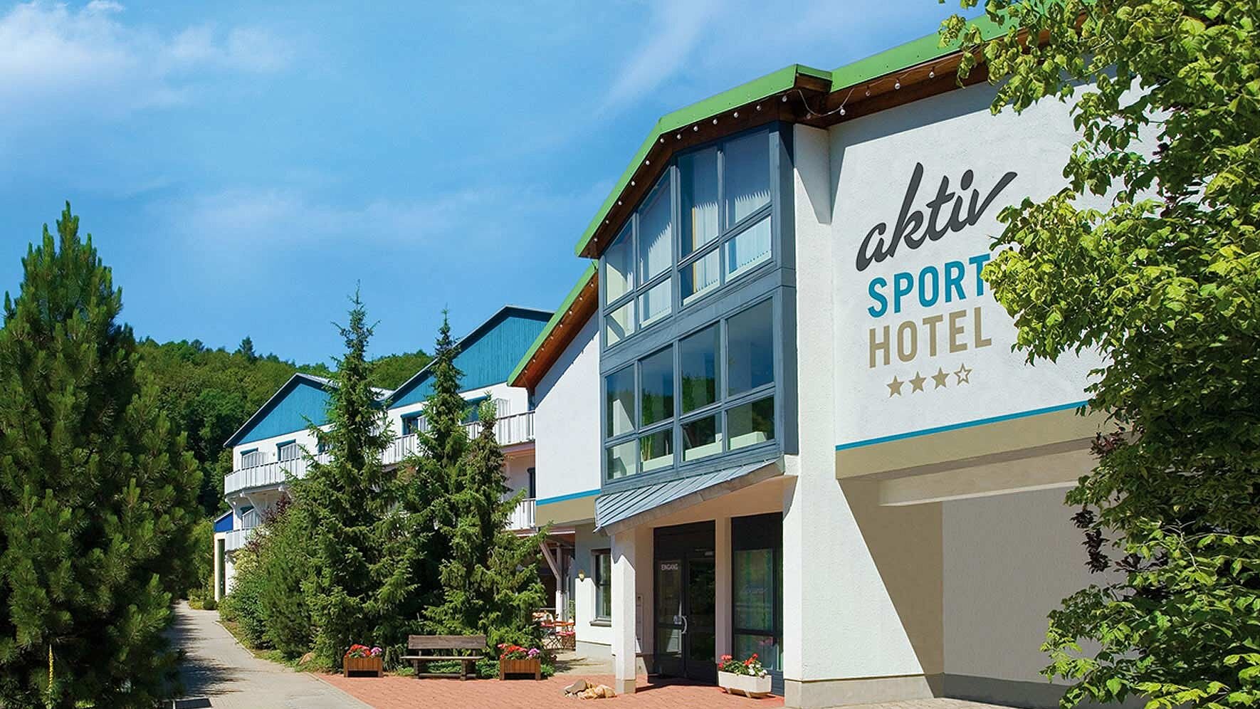 Außenansicht des Aktiv Sporthotels Pirna mit modernem Eingangsbereich, großen Fensterfronten und umliegenden Grünanlagen an einem sonnigen Tag.