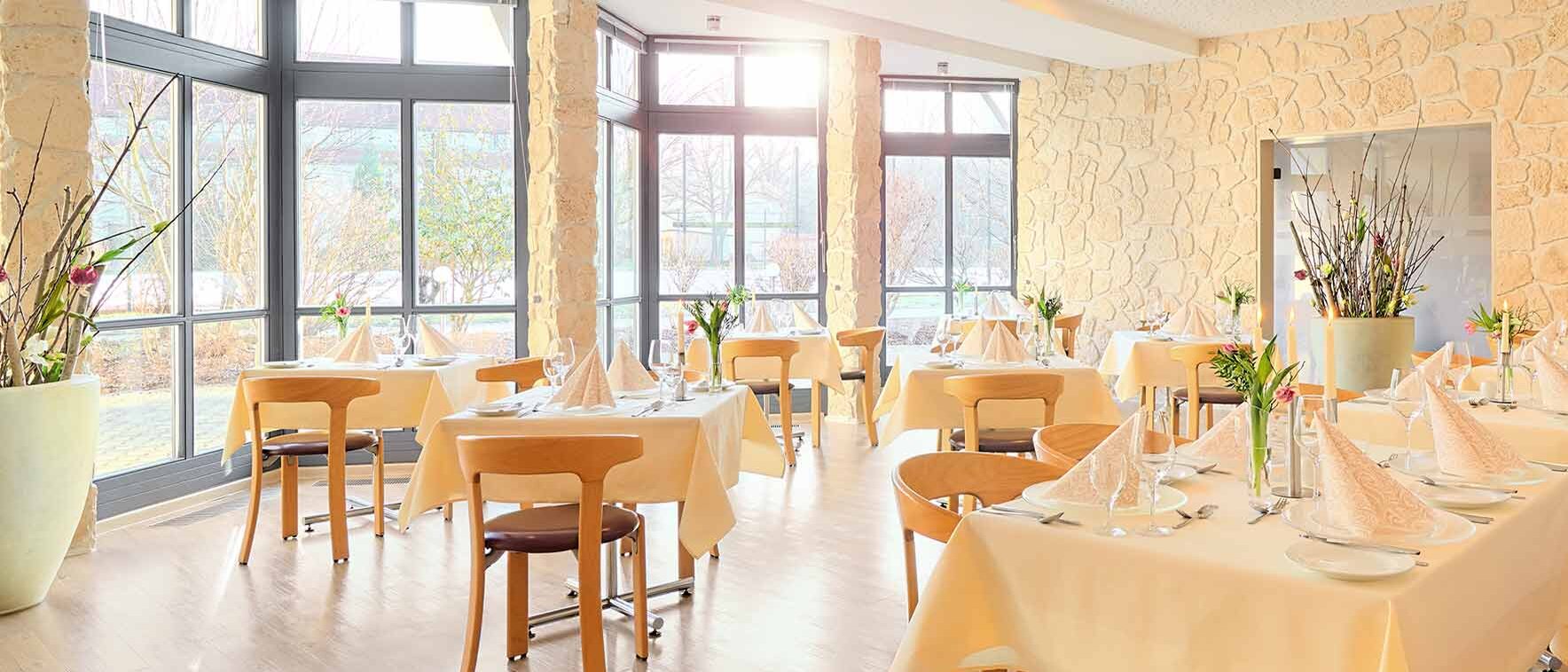 Das Foto präsentiert das lichtdurchflutete Restaurant des Aktiv Sporthotels Pirna und lädt mit großen Panoramafenstern und eleganten Natursteinwänden zu kulinarischen Highlights in stilvoller Atmosphäre ein. Die liebevoll eingedeckten Tische mit cremefarbenen Tischdecken, kunstvoll gefalteten Servietten und frischen Blumenarrangements verleihen dem Raum ein einladendes und modernen Flair – perfekt eingefangen auf diesem Bild.