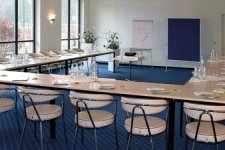 Heller Konferenzraum mit U-förmig angeordneten Tischen, gedeckt mit Notizblöcken, Stiften und Wasserflaschen. Mehrere Stühle stehen um die Tische. Im Hintergrund befinden sich ein Flipchart und eine Pinnwand. Große Fenster lassen viel Tageslicht in den Raum.