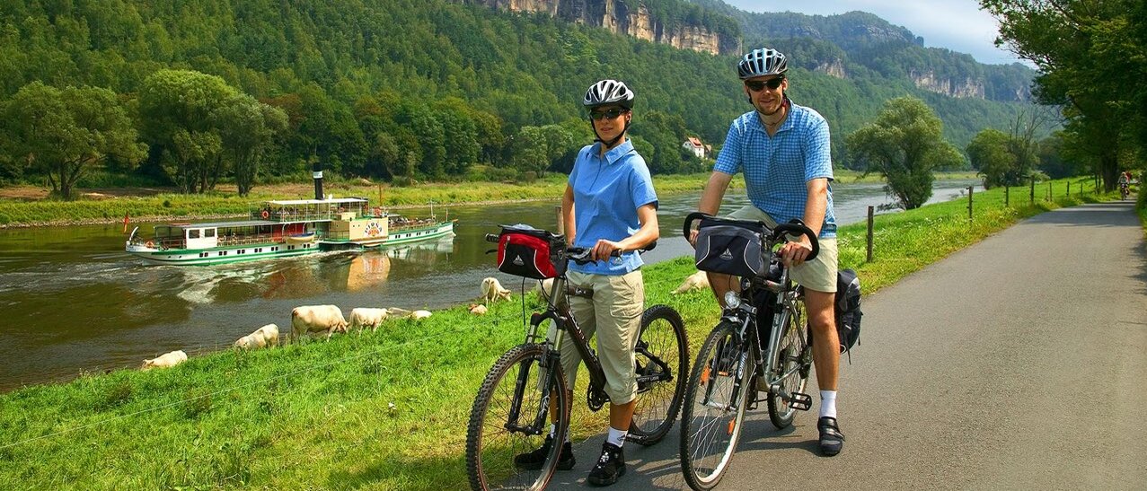 Zwei Personen stehen mit ihren Fahrrädern auf einem Radweg direkt an einem Fluss. Beide tragen Fahrradhelme und sportliche Kleidung. Auf dem Wasser fährt ein Ausflugsschiff vorbei, und im Hintergrund erheben sich die markanten Felsen der Sächsischen Schweiz. Die Szene wirkt sommerlich, aktiv und zeigt eine idyllische Fahrradtour inmitten einer beeindruckenden Landschaft.