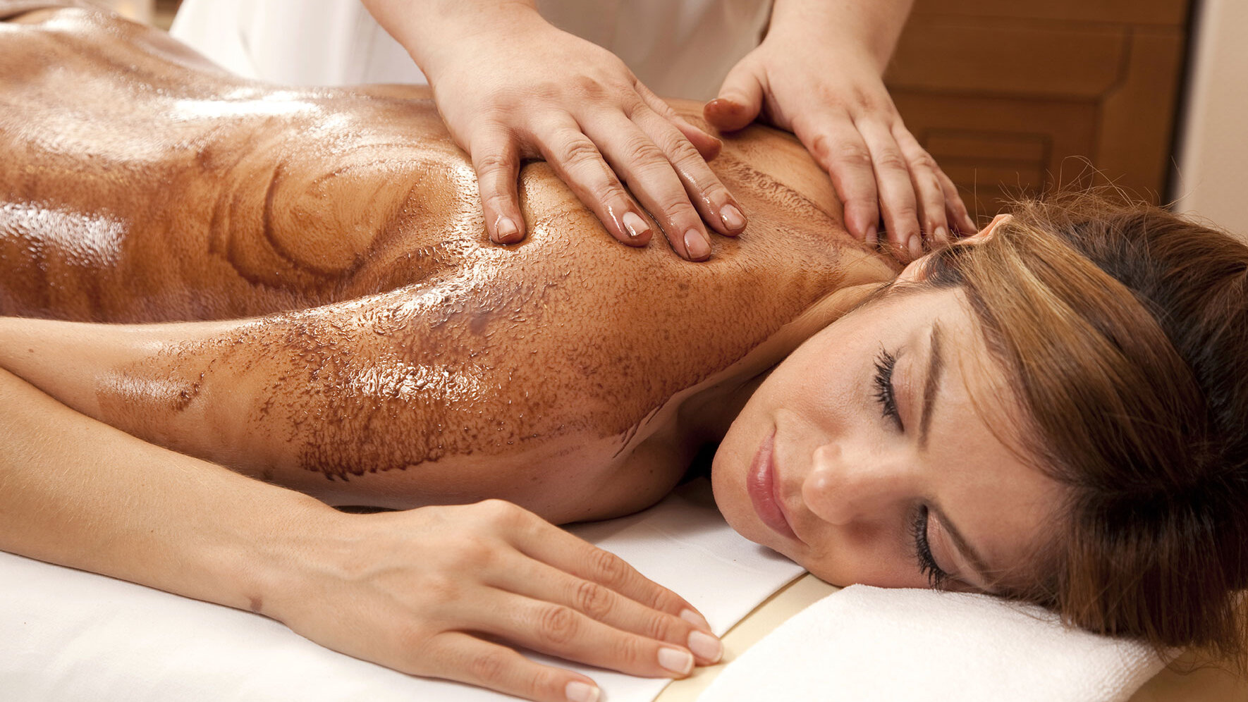 Eine Frau liegt entspannt auf einer Massageliege, während ihr Rücken mit warmem Öl eingerieben und massiert wird. Die Atmosphäre wirkt ruhig und wohltuend, mit sanfter Beleuchtung und einem warmen Farbton, wie man ihn aus Wellness- oder Spa-Bereichen kennt.