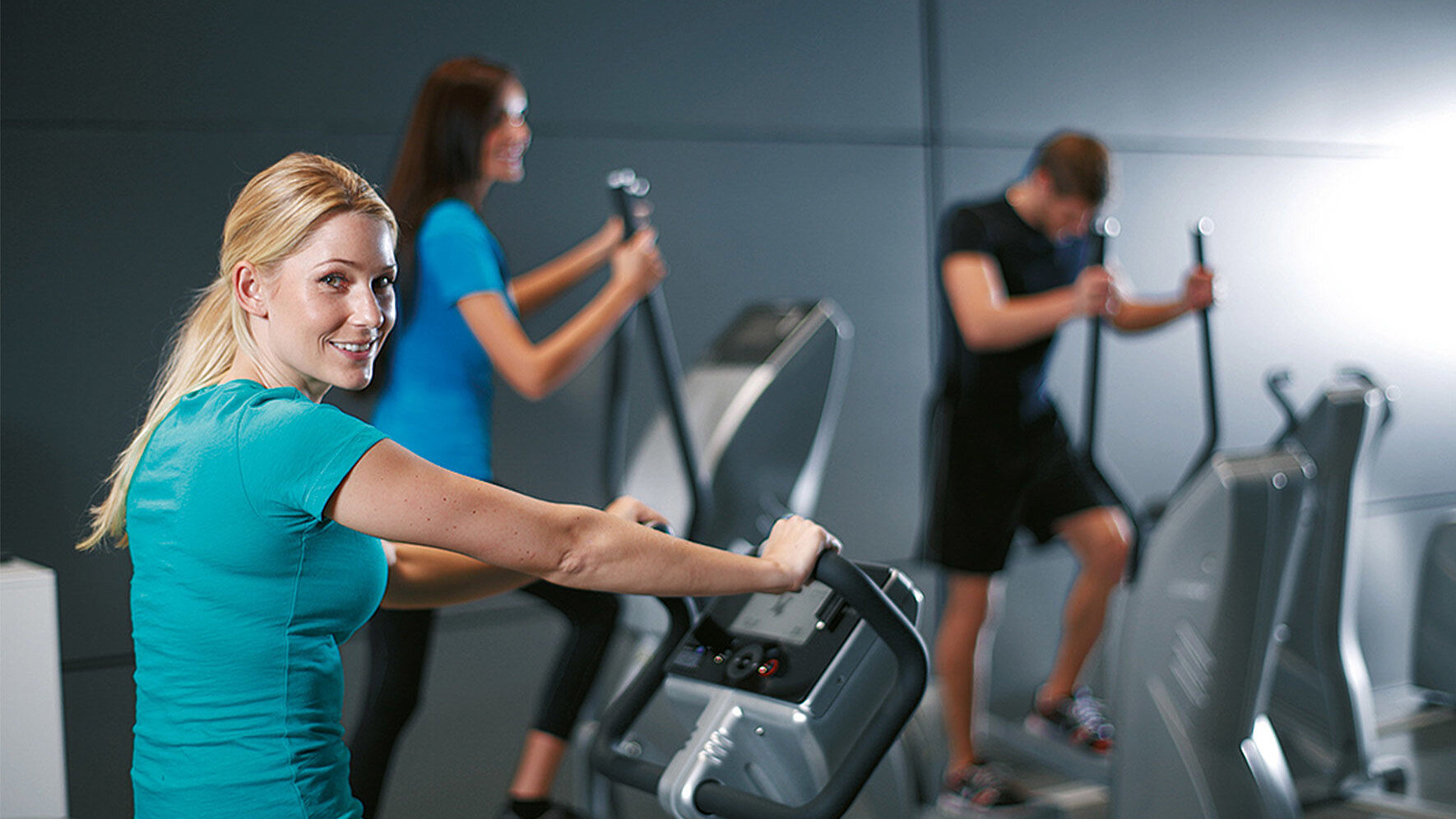 In unserem Fitnessstudio in Pirna trainieren drei Personen auf modernen Cardiogeräten. Im Vordergrund sitzt eine Frau in türkisfarbenem Sportshirt auf einem Ergometer und blickt lächelnd zur Kamera. Dahinter trainieren zwei weitere Personen auf Crosstrainern. Der Raum ist schlicht und grau gestaltet, gut beleuchtet und vermittelt eine aktive, sportliche Atmosphäre.