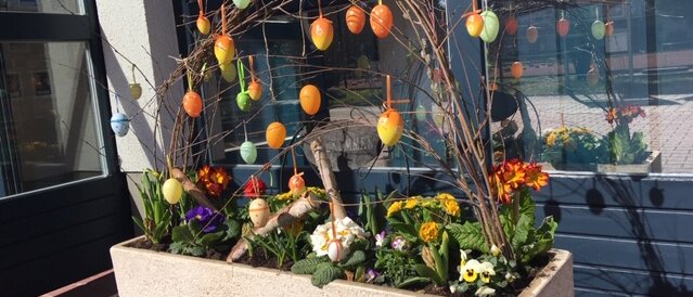 Ein großer rechteckiger Blumenkübel steht im Außenbereich vor dem Hotel. Darin blühen bunte Frühlingsblumen wie Stiefmütterchen und Primeln. Aus der Mitte ragen mehrere Zweige, an denen dekorativ bemalte Ostereier hängen. Das Arrangement wirkt frühlingshaft und festlich – passend zur Osterzeit.