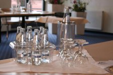 Nahaufnahme der bereitgestellten Erfrischungen auf dem Tagungstisch: In einem transparenten Halter stehen kleine Mineralwasserflaschen griffbereit neben polierten Gläsern und einem Flaschenkühler. Der helle, unscharfe Hintergrund mit blauem Teppich lässt die professionelle Raumausstattung erahnen.