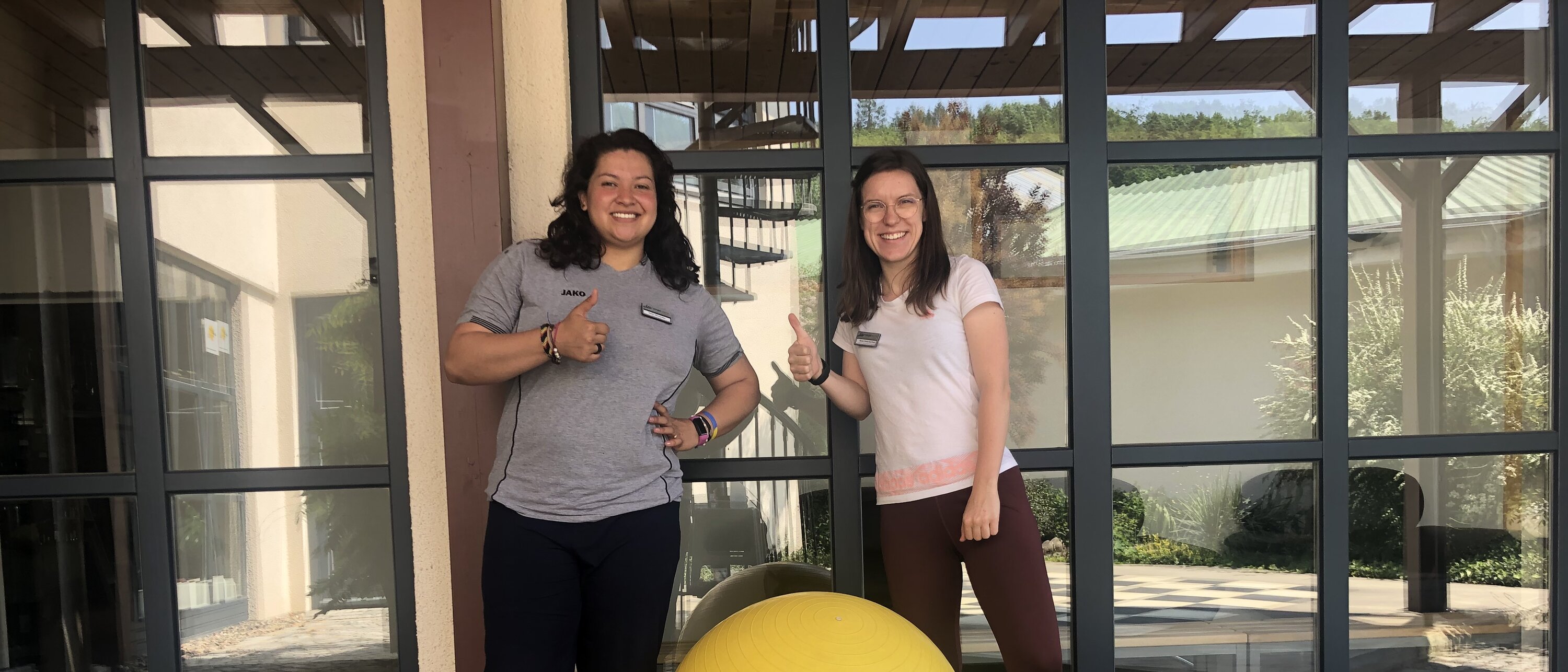 Zwei Frauen stehen vor dem Fitnessstudio und lächeln in die Kamera. Beide halten ein Getränk in der Hand. Vor ihnen steht ein großer, gelber Gymnastikball. Die Szene vermittelt gute Stimmung und Vorfreude auf einen aktiven Tag oder einen Kursmarathon.