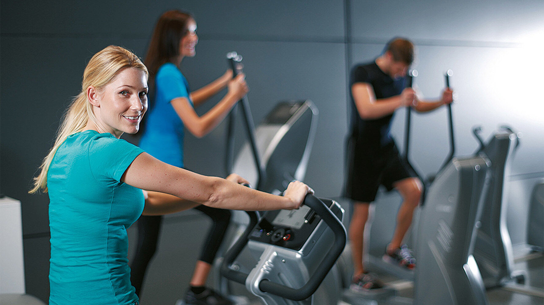 Das Foto zeigt das moderne Fitnessstudio im Aktiv Sporthotel: Drei motivierte Gäste trainieren konzentriert an hochwertigen Cardio-Geräten in lichtdurchfluteter und stilvoll gestalteter Umgebung. Die professionelle Ausstattung und die angenehme Atmosphäre lassen ideale Trainingsbedingungen erkennen – ein inspirierender Ort für sportliches Ambiente und aktive Erholung, wie in dieser Aufnahme eingefangen.