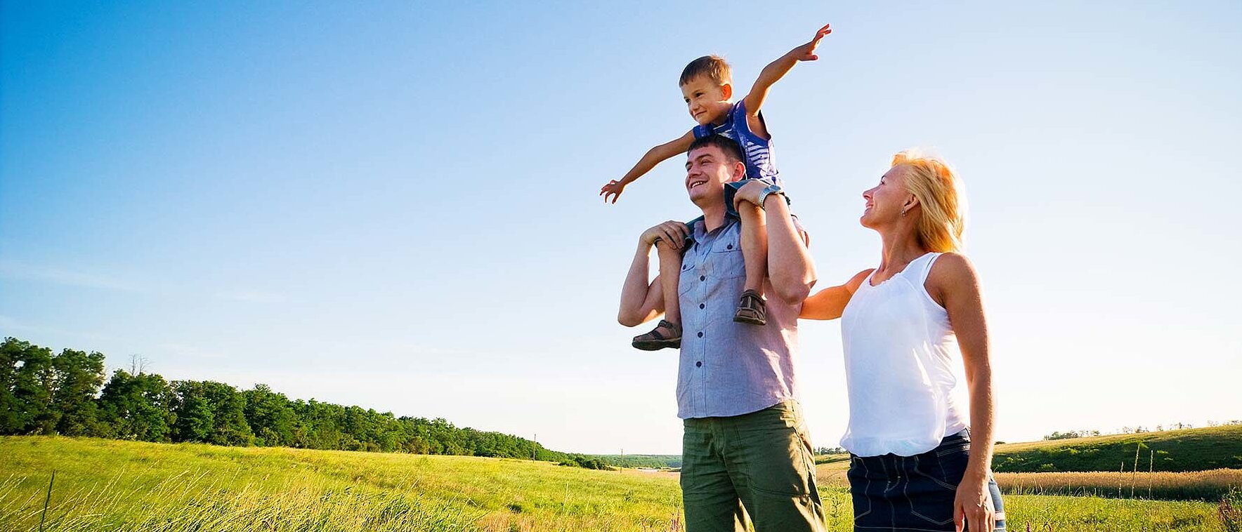 Eine fröhliche Familie steht auf einer weitläufigen grünen Wiese unter strahlend blauem Himmel. Ein Kind sitzt lachend auf den Schultern des Vaters und streckt spielerisch die Arme aus. Die Mutter steht daneben und schaut lächelnd zu. Das Bild vermittelt Leichtigkeit, Sommerstimmung und gemeinsame Aktivität – ideal für Sommerurlaub im Sporthotel.