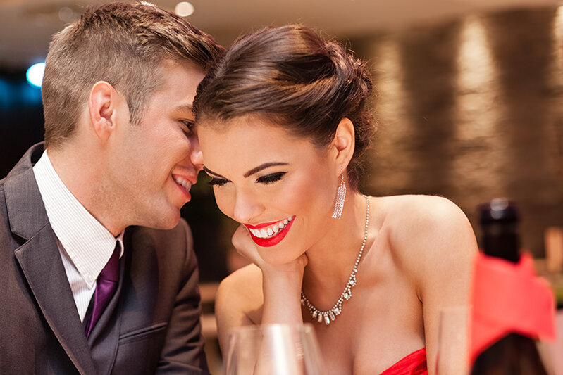 Ein elegant gekleidetes Paar sitzt eng beisammen in einem stimmungsvoll beleuchteten Restaurant. Der Mann flüstert der Frau etwas ins Ohr, während sie lächelnd und sichtbar erfreut reagiert. Die warme Atmosphäre, das Licht und die Nähe der beiden vermitteln romantische Stimmung – ideal für ein Valentinstagsdinner unter dem Motto ‚Love is in the Meal‘.