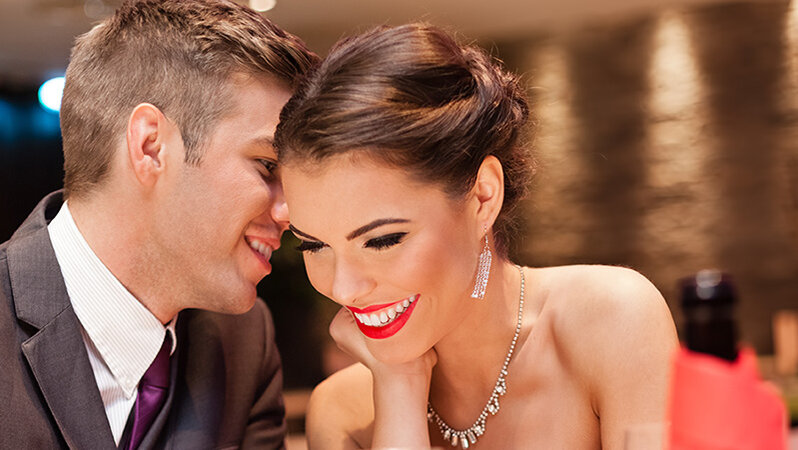 Ein elegant gekleidetes Paar sitzt eng beisammen in einem stimmungsvoll beleuchteten Restaurant. Der Mann flüstert der Frau etwas ins Ohr, während sie lächelnd und sichtbar erfreut reagiert. Die warme Atmosphäre, das Licht und die Nähe der beiden vermitteln romantische Stimmung – ideal für ein Valentinstagsdinner unter dem Motto ‚Love is in the Meal‘.