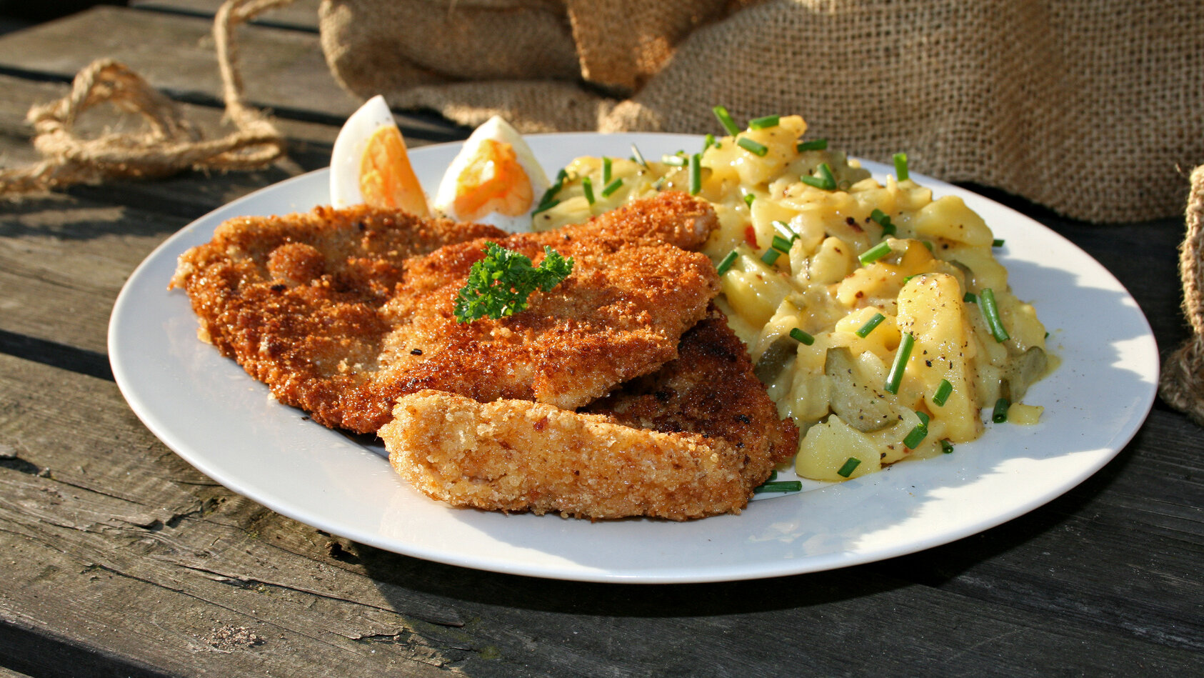 Auf einem weißen Teller liegt ein herzhaftes Gericht bestehend aus paniertem, goldbraun gebratenem Fleisch, Kartoffelbeilagen und würzigem Kohl- oder Krautsalat. Das Essen ist rustikal angerichtet und steht auf einem Holztisch im warmen Licht.