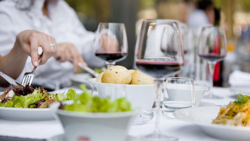 Eine Gruppe von Menschen sitzt an einem hell gedeckten Tisch im Freien und genießt verschiedene Gerichte. Auf dem Tisch stehen frische Salate, Pasta, Beilagen sowie mehrere Gläser Rotwein. Eine Person im Vordergrund schneidet ihr Essen. Die Szene vermittelt Genuss, Gastlichkeit und kulinarische Vielfalt – ideal als Motiv für kulinarische Wochen.