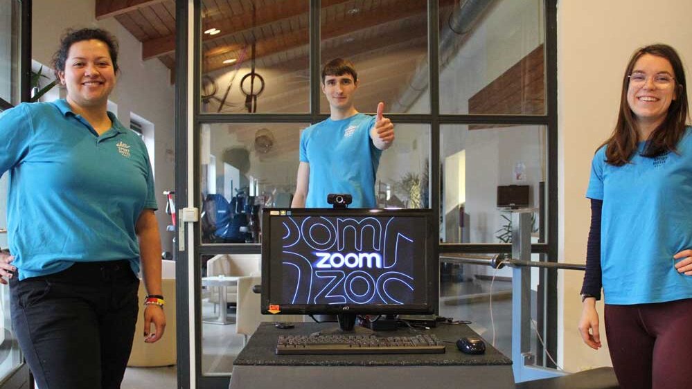 Drei Fitnesscoachs unseres Hotels stehen in Sportbekleidung in unserem modernen Fitnessbereich. Sie lächeln in die Kamera und präsentieren Trainingsgeräte wie einen Stepper und weitere Fitnessausstattung. Die Szene vermittelt Aktivität, Motivation und professionelle Betreuung.