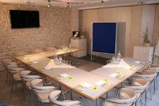 Blick in einen professionell ausgestatteten Tagungsraum mit Bestuhlung in U-Form. Der Raum besticht durch eine rustikale Natursteinwand mit montiertem Flachbildschirm. Die hellen Tische sind mit Tagungsgetränken, Schreibblöcken und Stiften arbeitsbereit vorbereitet. Im Präsentationsbereich stehen ein Rednerpult sowie eine blaue Pinnwand zur Verfügung.