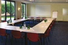 Heller, tageslichtdurchfluteter Tagungsraum mit bodentiefen Fenstern auf der linken Seite. Die Tische sind in klassischer U-Form gestellt, umgeben von roten Polsterstühlen auf einem dunkelblauen Teppichboden. Im Präsentationsbereich befinden sich eine fest installierte Leinwand und ein Flipchart. Auf den Tischen stehen Mineralwasser und Tagungsmaterialien bereit.