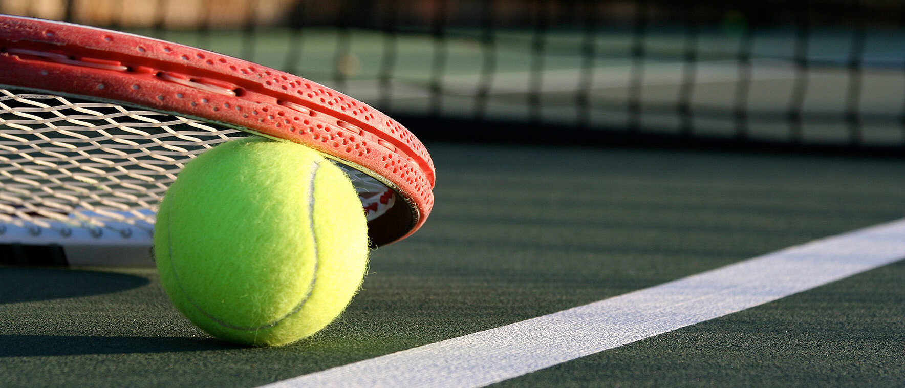 Ein gelber Tennisball liegt auf der weißen Linie eines Tennisplatzes direkt neben einem umgedrehten Tennisschläger. Im Hintergrund ist das Netz unscharf zu erkennen. Die Szene vermittelt sportliche Aktivität und die Atmosphäre eines professionellen Tennisplatzes.