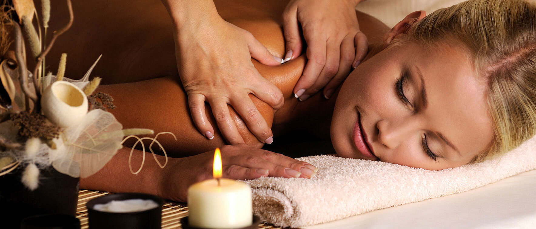 Eine Frau liegt entspannt auf dem Bauch auf einer Massagebank im Wellnessbereich des Hotels. Eine Masseurin massiert ihre Schultern und den oberen Rücken. Im Vordergrund stehen brennende Kerzen und dekorative Wellness-Elemente, die eine ruhige und wohltuende Atmosphäre schaffen.