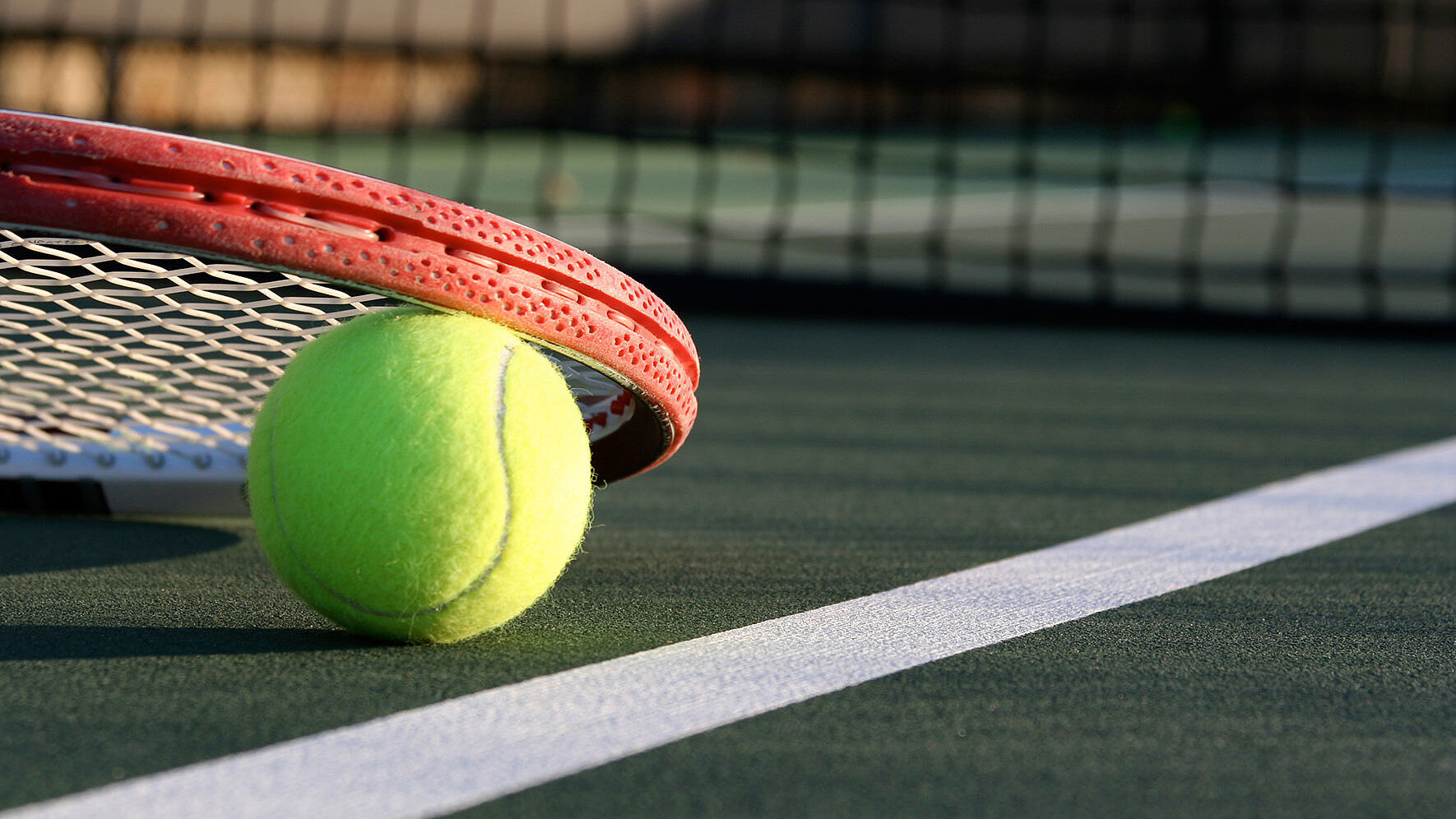 Ein gelber Tennisball liegt auf der weißen Linie eines Tennisplatzes direkt neben einem umgedrehten Tennisschläger. Im Hintergrund ist das Netz unscharf zu erkennen. Die Szene vermittelt sportliche Aktivität und die Atmosphäre eines professionellen Tennisplatzes.