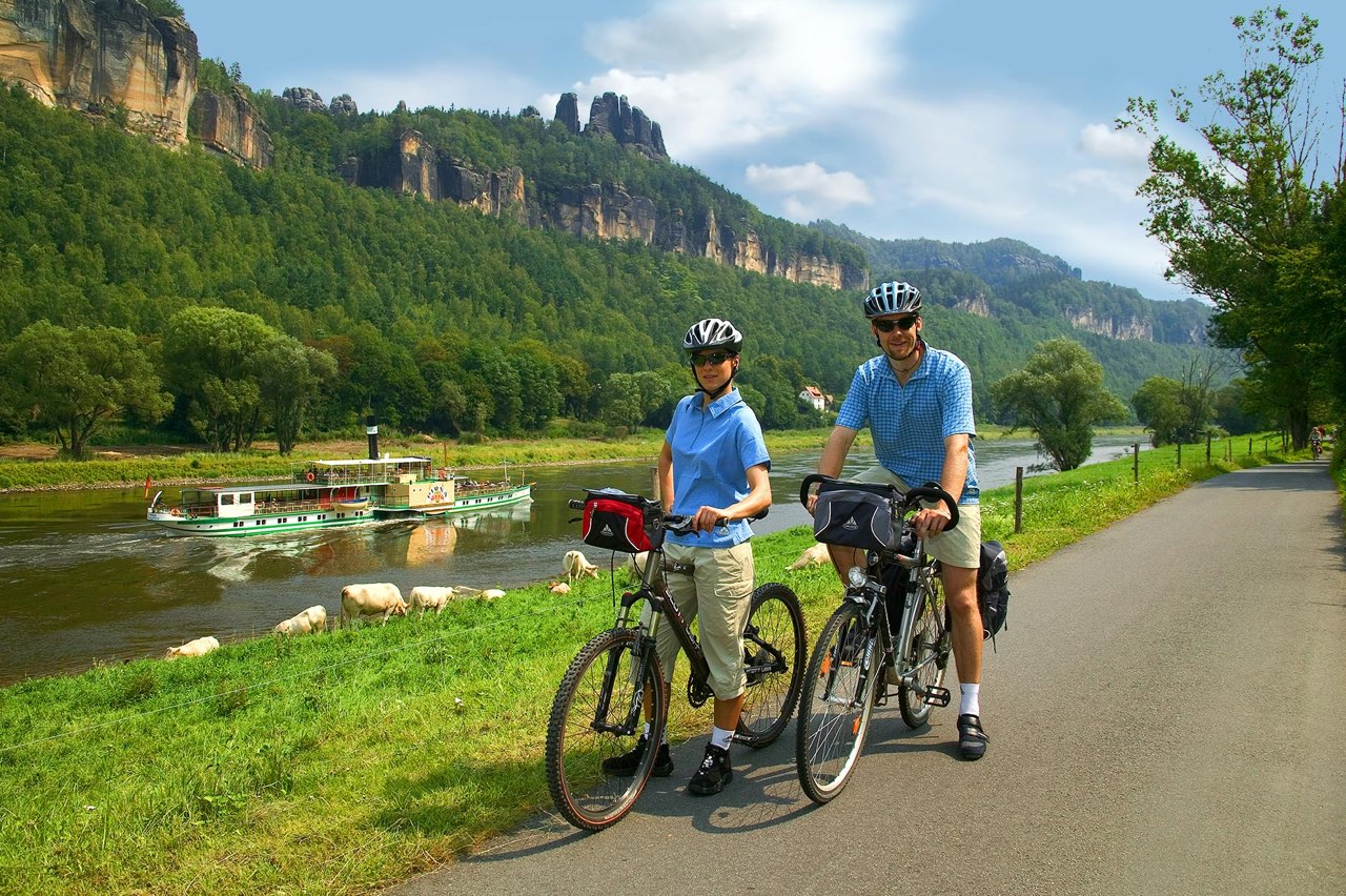 Zwei Personen stehen mit ihren Fahrrädern auf einem Radweg direkt an einem Fluss. Beide tragen Fahrradhelme und sportliche Kleidung. Auf dem Wasser fährt ein Ausflugsschiff vorbei, und im Hintergrund erheben sich die markanten Felsen der Sächsischen Schweiz. Die Szene wirkt sommerlich, aktiv und zeigt eine idyllische Fahrradtour inmitten einer beeindruckenden Landschaft.