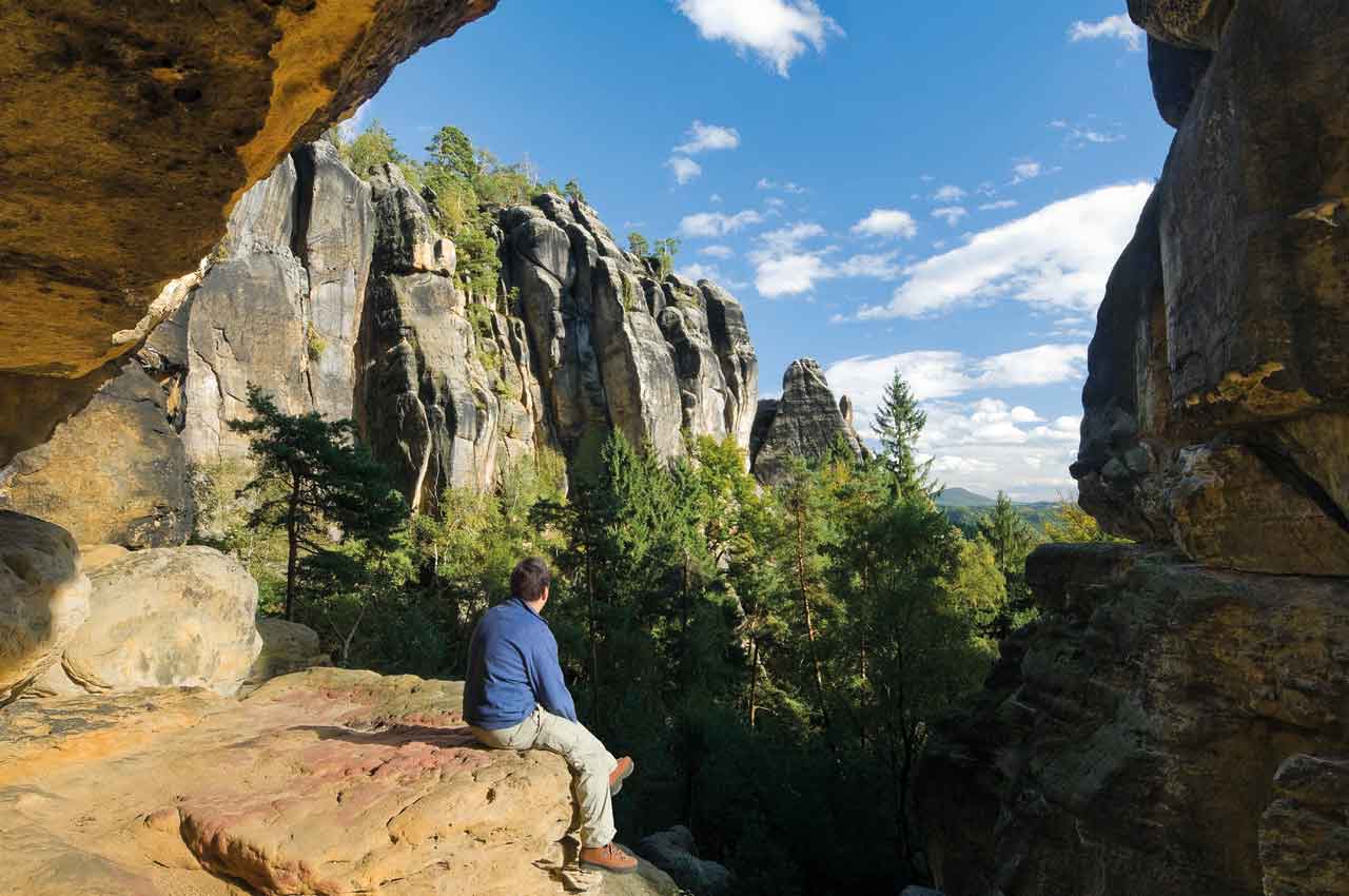 Eine Person sitzt auf einem Felsen und blickt über eine beeindruckende Felslandschaft mit hohen Sandsteinformationen und dichtem grünen Wald. Der Himmel ist blau mit einzelnen weißen Wolken. Die Szene vermittelt Ruhe, Naturverbundenheit und den Genuss einer Wanderpause inmitten der Sächsischen Schweiz.