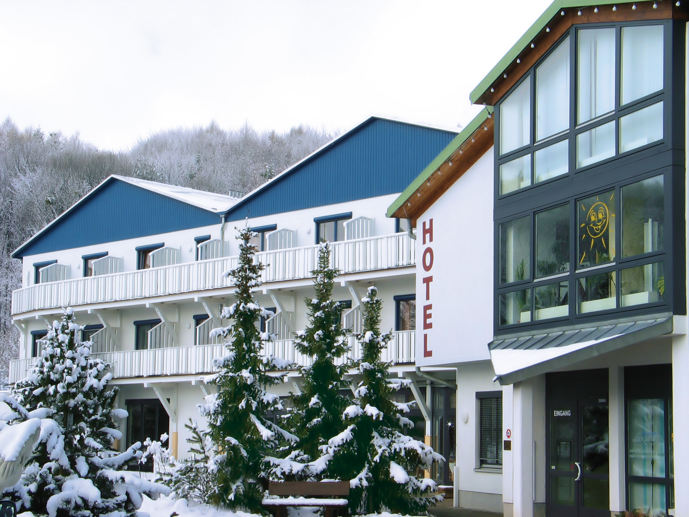 Auf dem Bild ist die Außenansicht des aktiv Sporthotel Pirna zu sehen. Es zeigt ein mehrstöckiges Gebäude mit weißer Fassade. Links befindet sich ein größerer Gebäudeteil mit blauen Schrägdächern und Balkonen. Rechts schließt ein moderner Gebäudeteil an, der große Fensterfronten besitzt. Vor dem Hotel stehen mehrere Tannenbäume, die leicht verschneit wirken. Die Szene vermittelt einen winterlichen, ruhigen Eindruck.