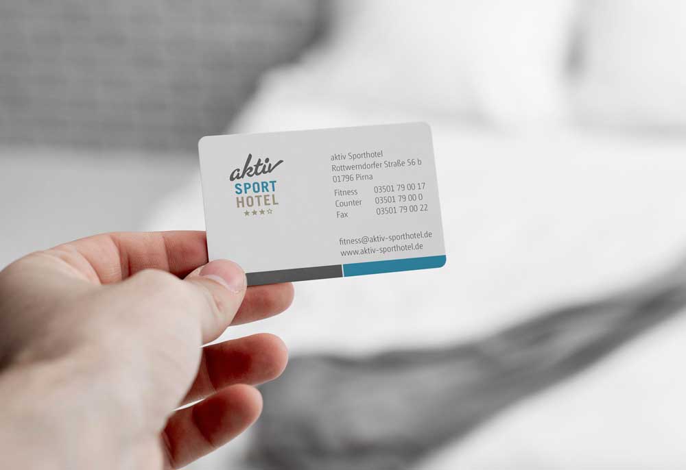 Eine Hand hält eine Business-Card des aktiv Sporthotels in die Kamera; im unscharfen Hintergrund ist ein Hotelbett mit weißer Bettwäsche zu erkennen.