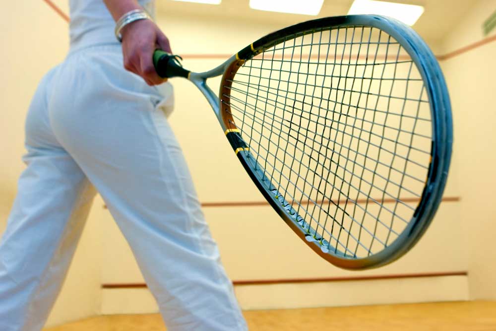 Nahaufnahme eines Squash-Spielers in weißer Hose, der einen Schläger in der Hand hält, stehend auf dem Holzboden eines Squash-Courts.