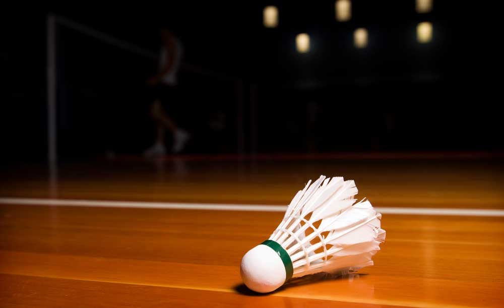 Badminton-Federball auf dem Hallenboden, im Hintergrund ein Spieler am Netz.