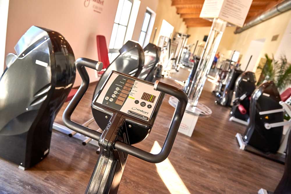 Auf dem Foto ist ein heller Trainingsraum eines Fitnessstudios zu sehen. Im Vordergrund steht ein ergonomisches Cardio-Gerät, vermutlich ein Fahrradergometer aus dem Milon-Zirkel. Man blickt direkt auf das digitale Display des Geräts, das verschiedene Tasten und Anzeigen besitzt. Die Form des Lenkers ist oval und umrahmt das Display.  Der Boden besteht aus dunklem Holz, was dem Raum eine warme Atmosphäre verleiht. Rechts und links vom Hauptgerät stehen weitere Kraft- und Ausdauergeräte, geordnet in einer Reihe, wie es typisch für einen Trainingszirkel ist.  Im Hintergrund fällt Tageslicht durch mehrere große, seitliche Fenster ins Studio. Die Decke ist aus holzbalkenähnlichen Elementen gefertigt, wodurch der Raum freundlich und offen wirkt. Insgesamt vermittelt das Bild den Eindruck eines modernen, gut beleuchteten Fitnessbereichs ohne Menschen.