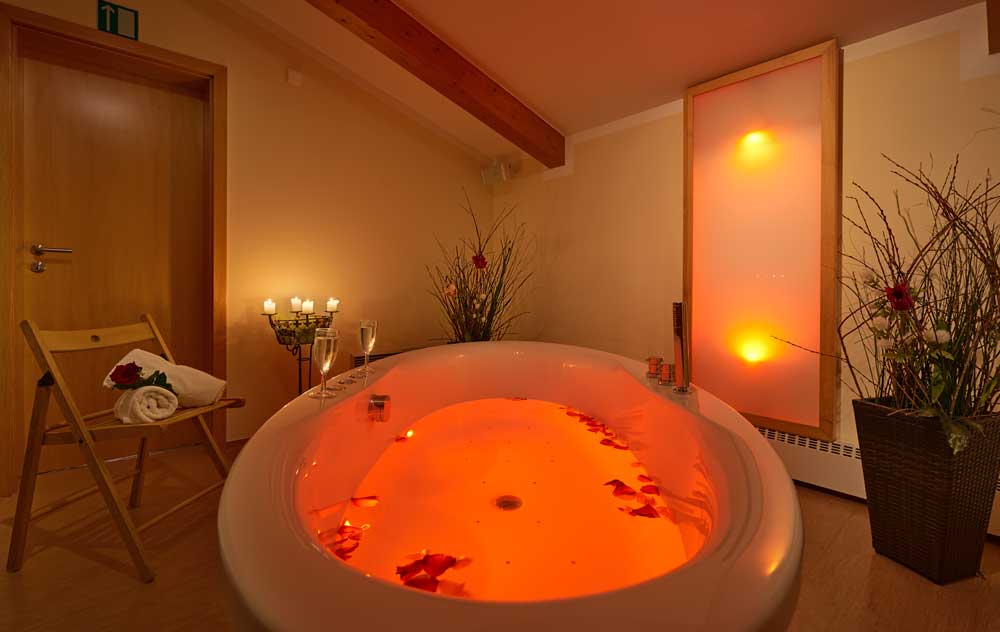 Stimmungsvoll beleuchteter Spa-Raum im Hotel mit einem warm orange leuchtenden Whirlpoolbad. Umgeben von Kerzen, Pflanzen und sanfter indirekter Beleuchtung entsteht eine ruhige, entspannende Wellness-Atmosphäre.