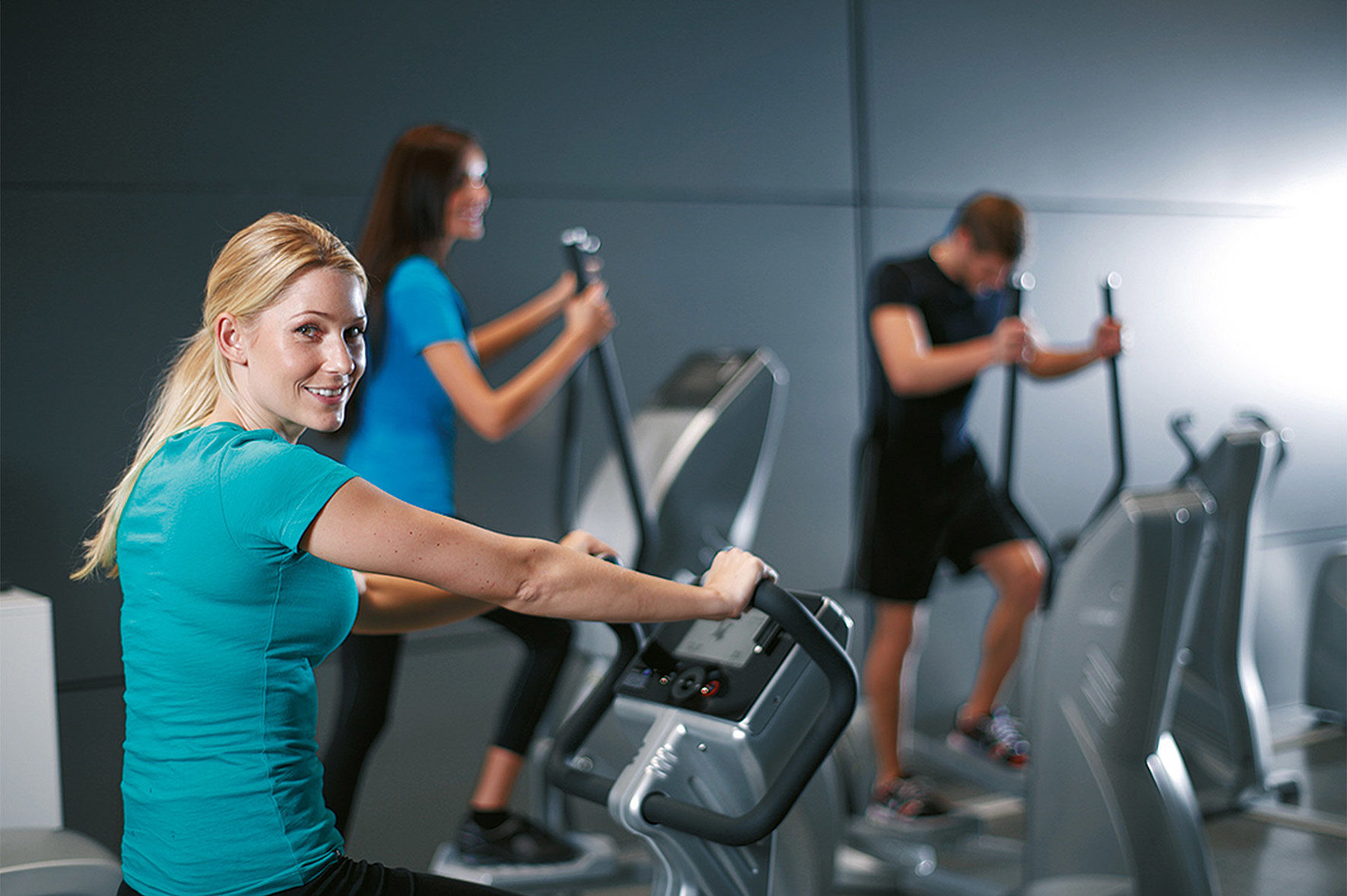 Das Foto zeigt das moderne Fitnessstudio im Aktiv Sporthotel Pirna: Drei motivierte Gäste trainieren konzentriert an hochwertigen Cardio-Geräten in lichtdurchfluteter und stilvoll gestalteter Umgebung. Die professionelle Ausstattung und die angenehme Atmosphäre lassen ideale Trainingsbedingungen erkennen – ein inspirierender Ort für sportliches Ambiente und aktive Erholung, wie in dieser Aufnahme eingefangen.