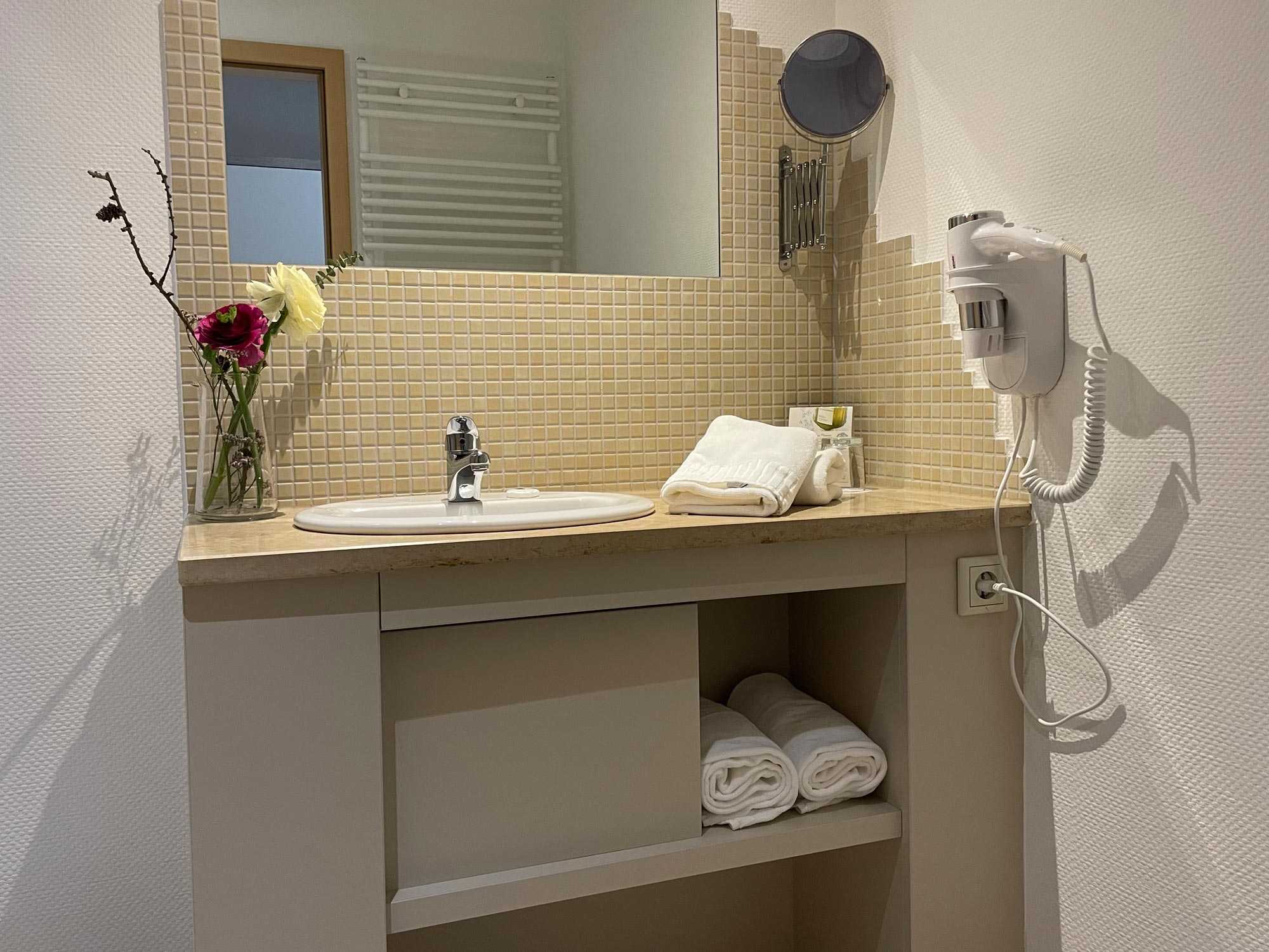 Blick auf den Waschtisch: Ein weißes Waschbecken vor beigen Mosaikfliesen, darüber ein Spiegel. Seitlich hängen ein Föhn und ein Kosmetikspiegel, frische Handtücher liegen bereit.