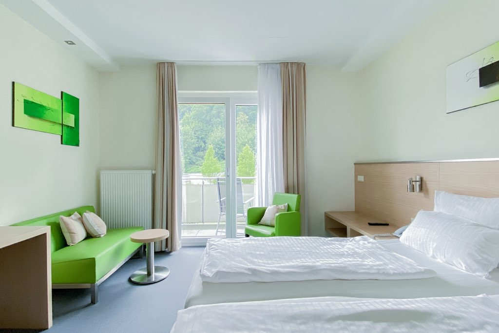 Gesamtansicht eines hellen Hotelzimmers: Rechts stehen die Betten, links befindet sich ein grünes Sofa mit passendem Sessel. Im Hintergrund führt eine Glastür auf den Balkon.