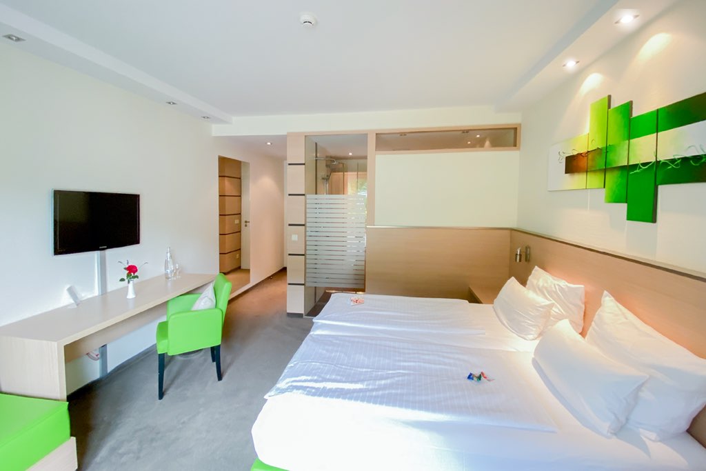 Weitwinkelansicht eines Superior-Hotelzimmers: Rechts das Bett mit grünem Wandbild, links ein langer Schreibtisch mit grünem Stuhl. Im Hintergrund trennt eine satinierte Glaswand den Schlafbereich vom Bad.