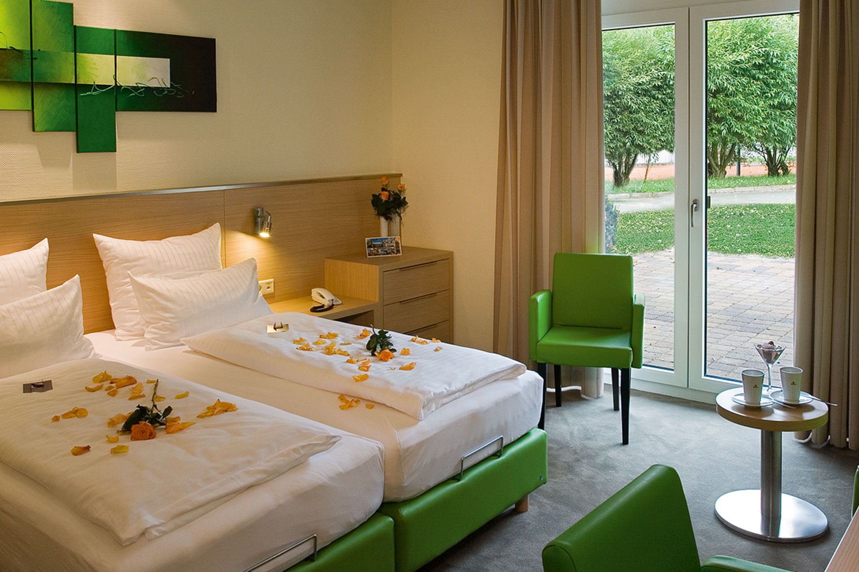 Modernes Hotelzimmer mit zwei einzelnen, frisch bezogenen Betten, dekoriert mit Blütenblättern. Grünes Mobiliar, ein kleiner Tisch mit Obst, große Fenster mit Blick in einen Garten und helle, freundliche Raumgestaltung.