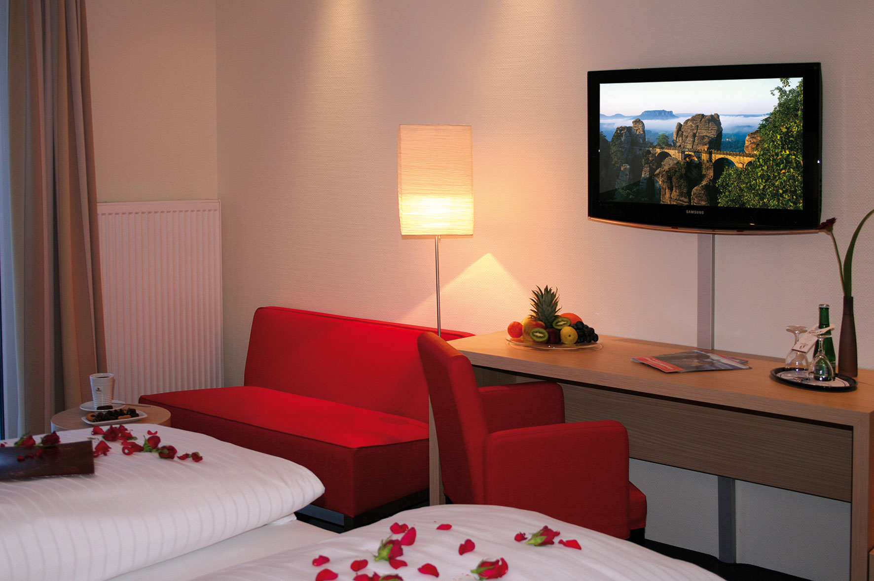 Romantischer Empfang im Hotelzimmer: Rosenblüten auf dem Bett, ein frischer Obstkorb und Getränke für einen besonderen Aufenthalt.