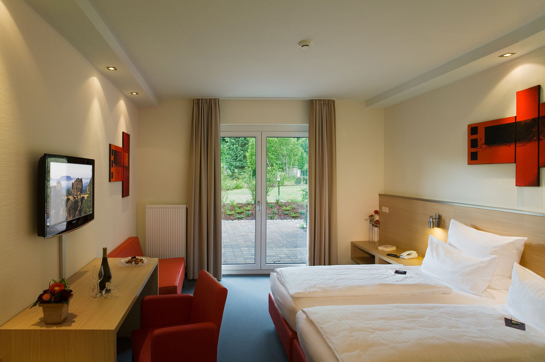 Blick in ein helles Hotelzimmer: Rechts stehen die Betten, links ein Schreibtisch mit Wand-TV und zwei roten Sesseln. Am Ende des Raumes führt eine Glastür ins Grüne.