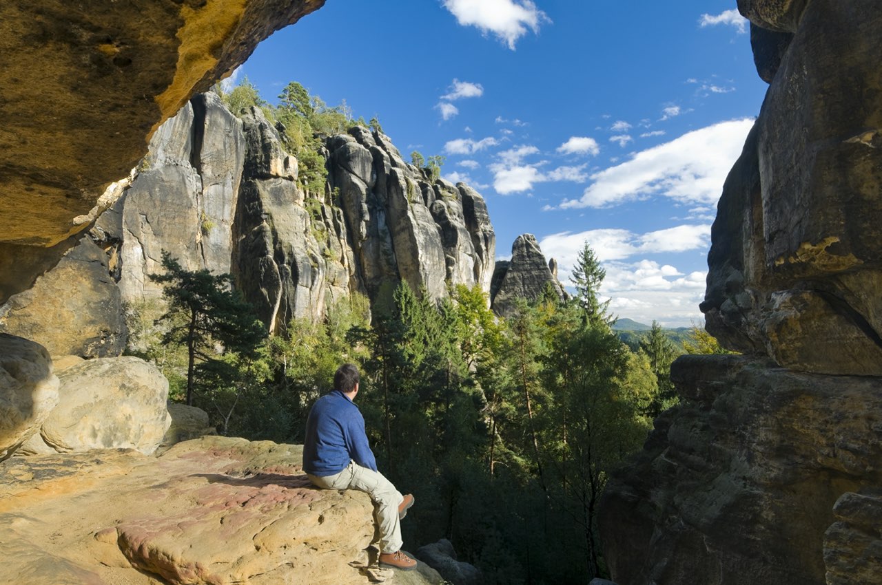 Spektakulärer Ausblick beim Wandern in der Sächsischen Schweiz: Ein Wanderer sitzt entspannt auf einem Felsvorsprung, eingerahmt von dunklen, massiven Felswänden einer Höhle oder Spalte. Der Blick geht hinaus auf die sonnenbeschienenen, zerklüfteten Sandsteinfelsen, grüne Baumwipfel und einen strahlend blauen Himmel.