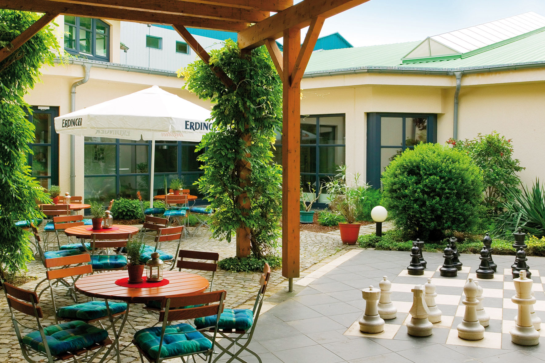 Gemütlicher Außenbereich unseres Restaurants mit grünen Pflanzen, Holztischen und Stühlen, schattenspendenden Pergolen und einem großen Outdoor-Schachspiel auf einer Terrasse.