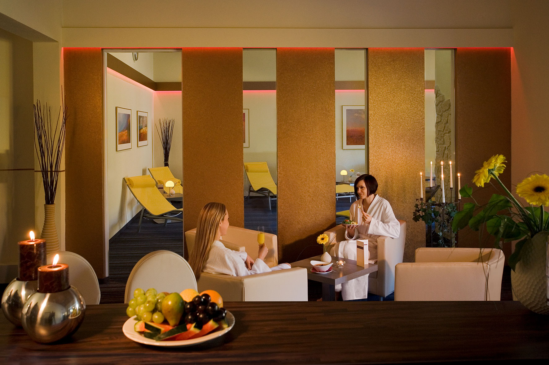 Wellnessbereich mit sanfter Beleuchtung: Zwei Personen in weißen Bademänteln sitzen an einem kleinen Tisch, umgeben von bequemen Liegen, warmen Farben, Kerzen, Blumen und einer Schale mit frischem Obst im Vordergrund.