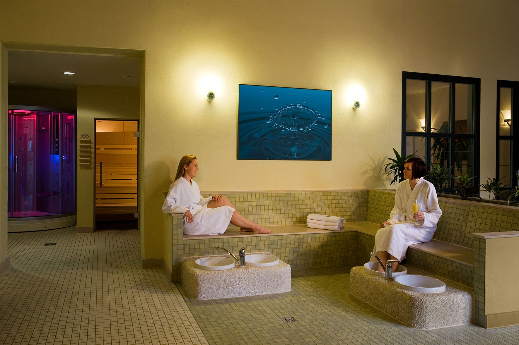 Atmosphärischer Wellnessbereich mit Mosaik-Design: Zwei Frauen entspannen in Bademänteln auf einer gemauerten Sitzbank. Davor stehen Fußbecken für wohltuende Wechselbäder bereit. Links im Hintergrund sieht man den Zugang zur Holz-Sauna sowie eine moderne Erlebnisdusche mit stimmungsvollem, farbigem Licht (Pink/Blau).