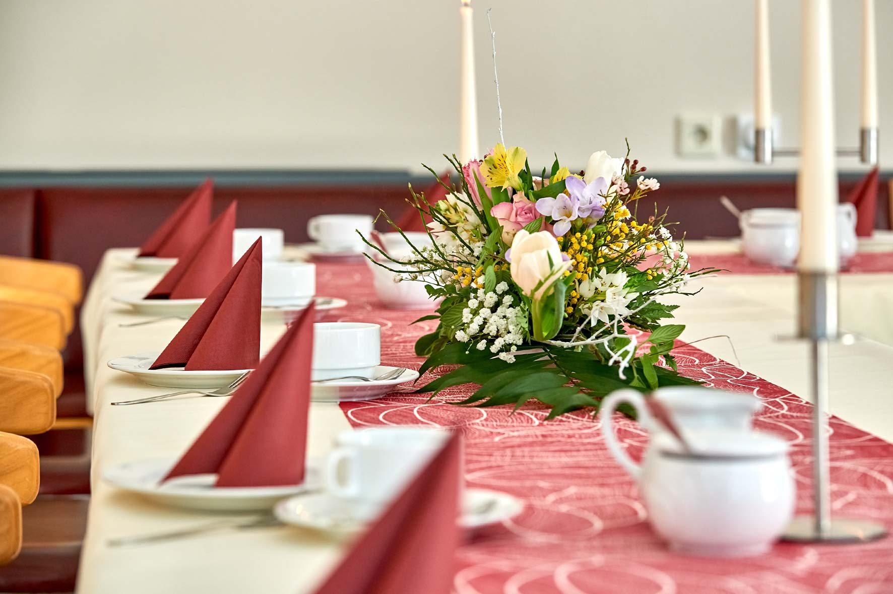 Festlich gedeckter Tisch in einem Veranstaltungsraum. Auf der langen Tafel liegen ein rotes Tischband mit Muster, darauf ein großer Blumenstrauß aus weißen, rosa und gelben Blüten. Entlang der Tischmitte stehen hohe weiße Kerzen. Die Plätze sind mit weißen Tassen, Untertellern, kleinen Kännchen sowie rot gefalteten Stoffservietten eingedeckt. Im Hintergrund sind weitere eingedeckte Plätze zu erkennen.
