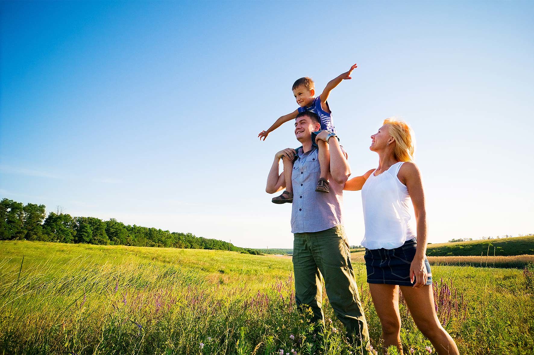 Eine fröhliche Familie steht auf einer weitläufigen grünen Wiese unter strahlend blauem Himmel. Ein Kind sitzt lachend auf den Schultern des Vaters und streckt spielerisch die Arme aus. Die Mutter steht daneben und schaut lächelnd zu. Das Bild vermittelt Leichtigkeit, Sommerstimmung und gemeinsame Aktivität – ideal für Sommerurlaub im Sporthotel.