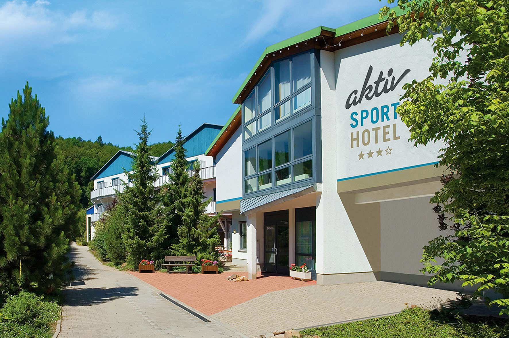 Außenansicht des Aktiv Sporthotels Pirna mit modernem Eingangsbereich, großen Fensterfronten und umliegenden Grünanlagen an einem sonnigen Tag.