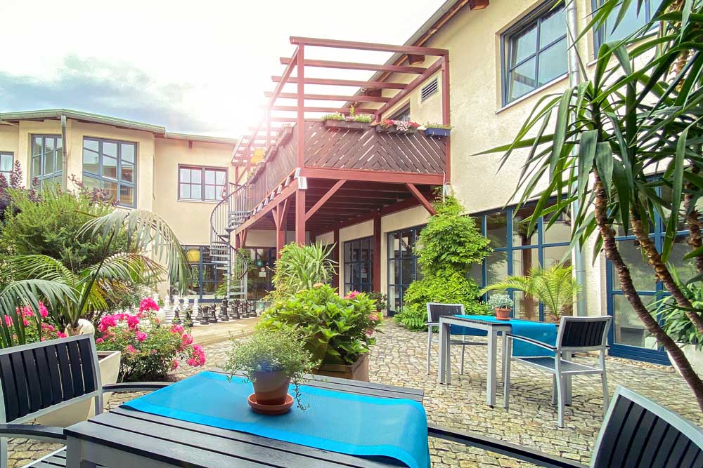 Auf dem Bild sieht man den liebevoll gestalteten Außenbereich des Wellness- und Spa-Bereichs im Aktiv Sporthotel Pirna. Zwischen üppigem Grün, blühenden Pflanzen und mediterranen Gestaltungselementen stehen moderne Sitzgruppen mit blauen Tischdecken bereit – ideal zum Entspannen an der frischen Luft. Die warme Fassade des Hotels und der hölzerne Balkon verleihen dem Innenhof eine behagliche, fast mediterrane Atmosphäre. Das Zusammenspiel aus Licht, Natur und gemütlicher Einrichtung unterstreicht den erholsamen Charakter des Wellnesshotels in der Sächsischen Schweiz.