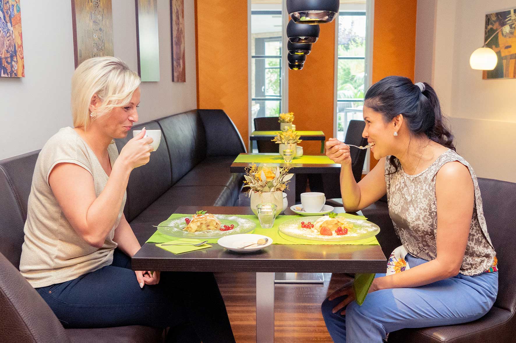 Zwei Frauen sitzen in einer modern gestalteten Lounge an einem gemütlichen Tisch, genießen gemeinsam ein Frühstück und unterhalten sich. Der Raum ist hell, freundlich dekoriert und wirkt einladend.